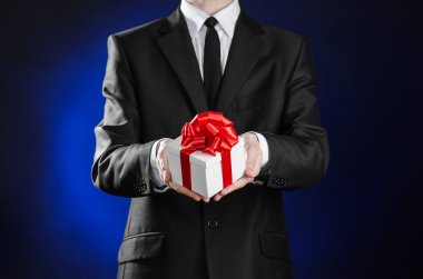 Theme holidays and gifts: a man in a black suit holds an exclusive gift in a white box wrapped with red ribbon and bow on a dark blue background in studio