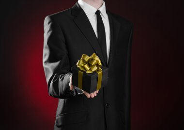 Theme holidays and gifts: a man in a black suit holds exclusive gift wrapped in a black box with gold ribbon and bow on a dark red background in studio
