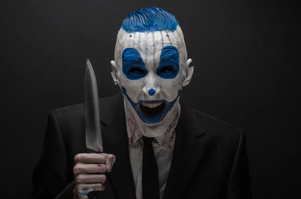 Terrible clown and Halloween theme: Crazy blue clown in a black suit with a knife in his hand isolated on a dark background in the studio — 图库照片