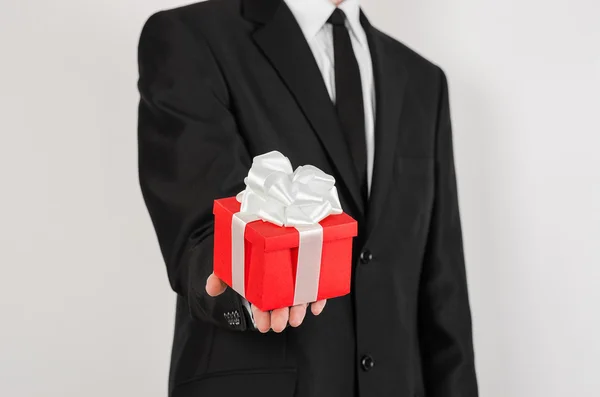 Férias temáticas e presentes: um homem de terno preto detém presente exclusivo embrulhado em caixa vermelha com fita branca e arco isolado em um fundo branco no estúdio — Fotografia de Stock