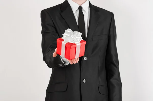 Theme holidays and gifts: a man in a black suit holds exclusive gift wrapped in red box with white ribbon and bow isolated on a white background in studio — Zdjęcie stockowe