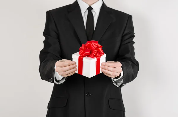 Theme holidays and gifts: a man in a black suit holds an exclusive gift in a white box wrapped with red ribbon and bow isolated on a white background in studio — Stockfoto