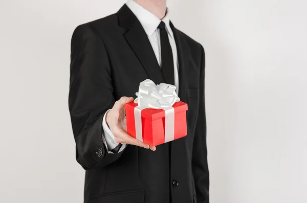 Vacaciones temáticas y regalos: un hombre en traje negro sostiene regalo exclusivo envuelto en caja roja con cinta blanca y lazo aislado sobre un fondo blanco en el estudio —  Fotos de Stock