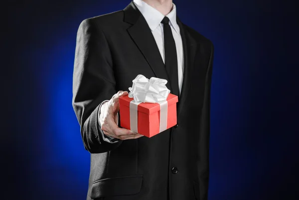 Theme holidays and gifts: a man in a black suit holds exclusive gift wrapped in red box with white ribbon and bow on a dark blue background in studio — 스톡 사진