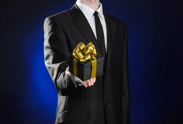 Férias temáticas e presentes: um homem em um terno preto detém presente exclusivo envolto em uma caixa preta com fita de ouro e arco em um fundo azul escuro em estúdio — Fotografia de Stock