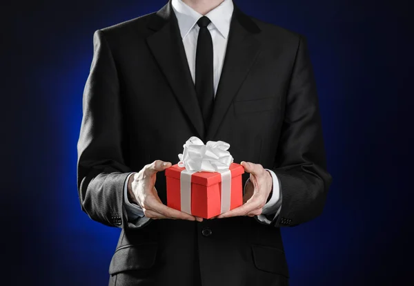 Vacaciones temáticas y regalos: un hombre con un traje negro sostiene regalo exclusivo envuelto en caja roja con cinta blanca y lazo sobre un fondo azul oscuro en el estudio —  Fotos de Stock