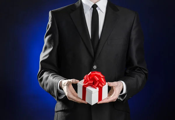 Férias temáticas e presentes: um homem de terno preto detém um presente exclusivo em uma caixa branca envolta com fita vermelha e arco em um fundo azul escuro em estúdio — Fotografia de Stock