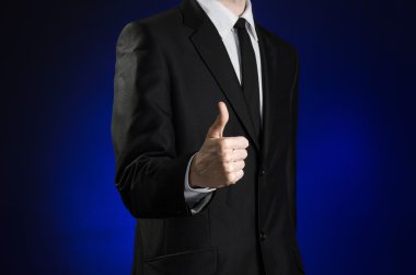Businessman and gesture topic: a man in a black suit and white shirt showing a thumbs up hand on a dark blue background in studio isolated