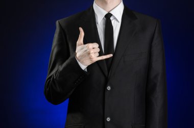 Businessman and gesture topic: a man in a black suit and white shirt showing sign a telephone hands on a dark blue background in studio isolated