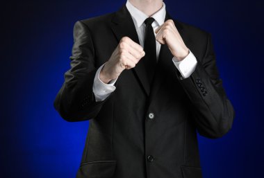 Businessman and gesture topic: a man in a black suit and white shirt holding his fists in front of him on a dark blue background in studio isolated