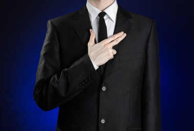 Businessman and gesture topic: a man in a black suit and white shirt, put his hand on his chest as a sign of a pistol on a dark blue background in studio isolated