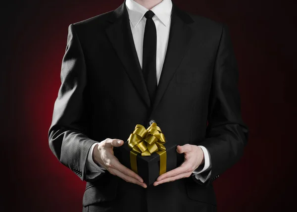 Theme holidays and gifts: a man in a black suit holds exclusive gift wrapped in a black box with gold ribbon and bow on a dark red background in studio — Stok fotoğraf