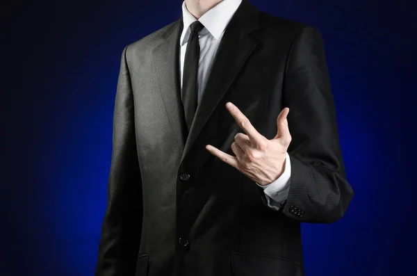 Businessman and gesture topic: a man in a black suit and white shirt shows a hand sign rock on a dark blue background in studio isolated — Stockfoto