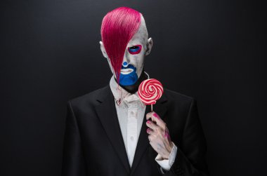 Clown and Halloween theme: Scary clown with pink hair in a black jacket with candy in hand on a dark background in the studio