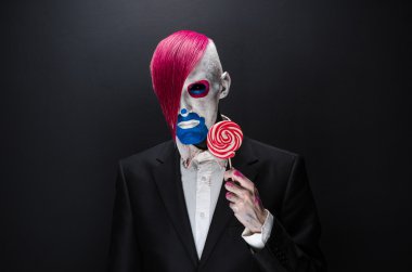 Clown and Halloween theme: Scary clown with pink hair in a black jacket with candy in hand on a dark background in the studio
