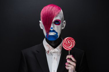 Clown and Halloween theme: Scary clown with pink hair in a black jacket with candy in hand on a dark background in the studio