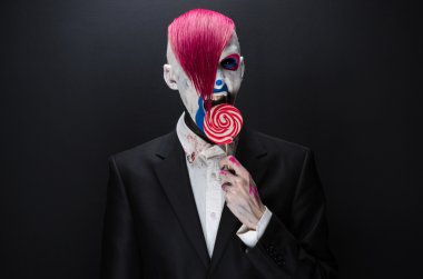 Clown and Halloween theme: Scary clown with pink hair in a black jacket with candy in hand on a dark background in the studio