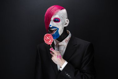 Clown and Halloween theme: Scary clown with pink hair in a black jacket with candy in hand on a dark background in the studio
