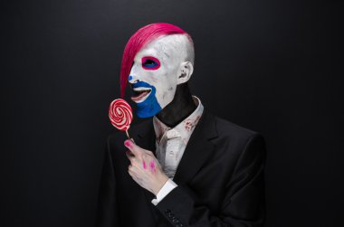 Clown and Halloween theme: Scary clown with pink hair in a black jacket with candy in hand on a dark background in the studio