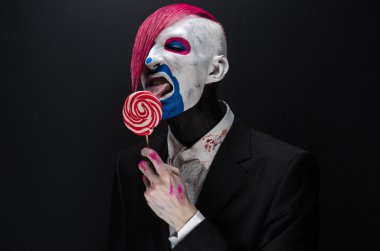 Clown and Halloween theme: Scary clown with pink hair in a black jacket with candy in hand on a dark background in the studio