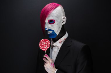 Clown and Halloween theme: Scary clown with pink hair in a black jacket with candy in hand on a dark background in the studio