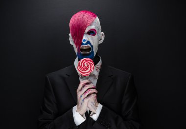 Clown and Halloween theme: Scary clown with pink hair in a black jacket with candy in hand on a dark background in the studio