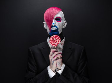 Clown and Halloween theme: Scary clown with pink hair in a black jacket with candy in hand on a dark background in the studio