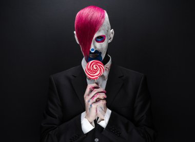 Clown and Halloween theme: Scary clown with pink hair in a black jacket with candy in hand on a dark background in the studio