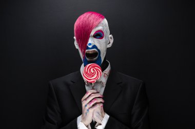 Clown and Halloween theme: Scary clown with pink hair in a black jacket with candy in hand on a dark background in the studio