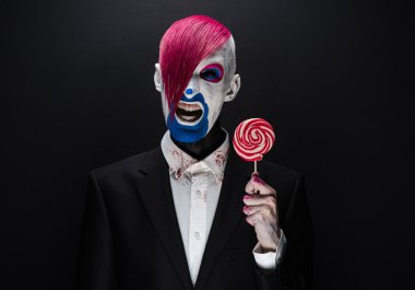 Clown and Halloween theme: Scary clown with pink hair in a black jacket with candy in hand on a dark background in the studio