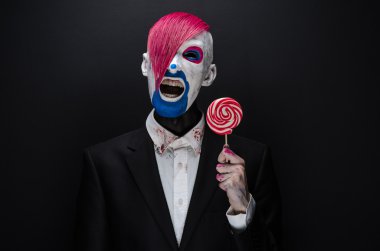 Clown and Halloween theme: Scary clown with pink hair in a black jacket with candy in hand on a dark background in the studio