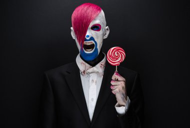 Clown and Halloween theme: Scary clown with pink hair in a black jacket with candy in hand on a dark background in the studio