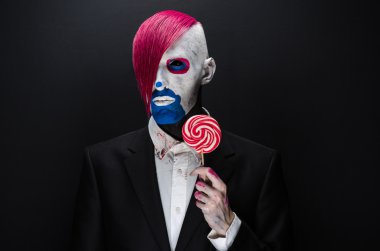 Clown and Halloween theme: Scary clown with pink hair in a black jacket with candy in hand on a dark background in the studio