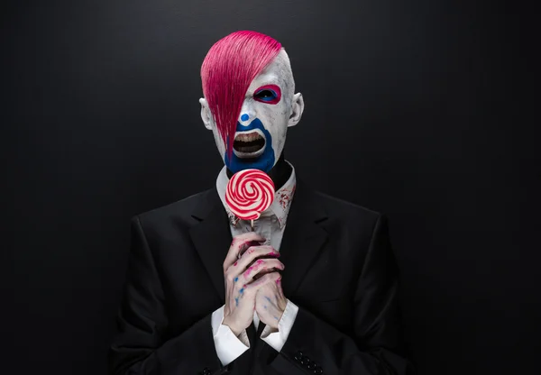 Clown e tema Halloween: clown spaventoso con i capelli rosa in una giacca nera con caramelle in mano su uno sfondo scuro in studio — Foto Stock