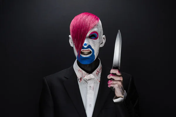 Clown and Halloween theme: Scary clown with pink hair in a black suit with a knife in his hand on a dark background in the studio — Stok fotoğraf