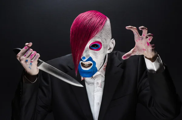 Clown and Halloween theme: Scary clown with pink hair in a black suit with a knife in his hand on a dark background in the studio — Stok fotoğraf