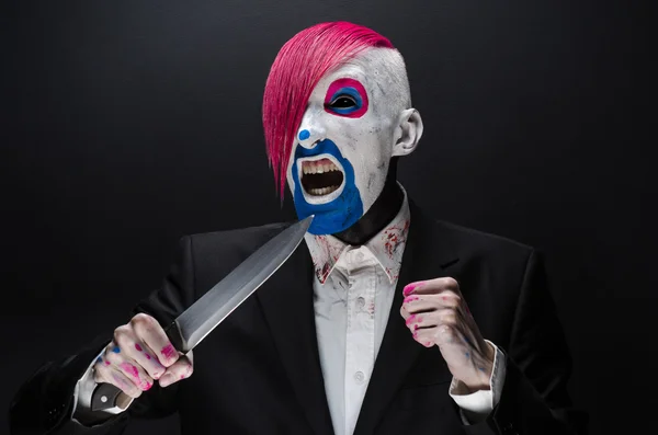 Clown and Halloween theme: Scary clown with pink hair in a black suit with a knife in his hand on a dark background in the studio — Stok fotoğraf