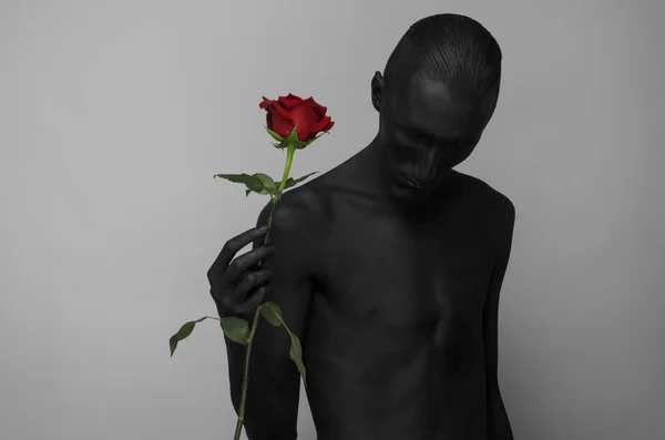 Tema gotico e Halloween: un uomo con la pelle nera che tiene una rosa rossa, la morte nera isolata su uno sfondo grigio in studio — Foto Stock