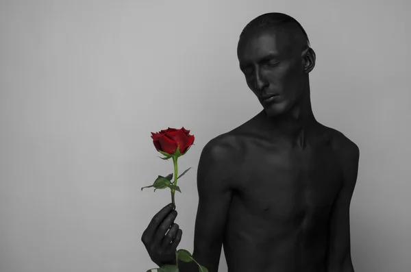 Tema gotico e Halloween: un uomo con la pelle nera che tiene una rosa rossa, la morte nera isolata su uno sfondo grigio in studio — Foto Stock