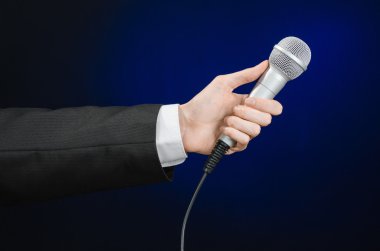 Business and speech topic: Man in black suit holding a microphone on a gray dark blue isolated background in studio