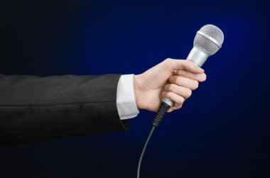 Business and speech topic: Man in black suit holding a microphone on a gray dark blue isolated background in studio