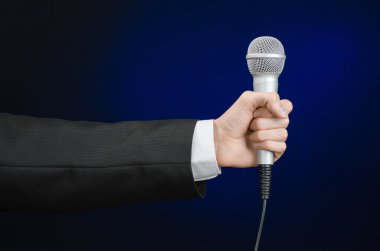 Business and speech topic: Man in black suit holding a microphone on a gray dark blue isolated background in studio