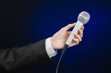 Business and speech topic: Man in black suit holding a microphone on a gray dark blue isolated background in studio