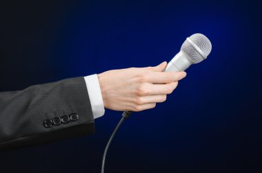 Business and speech topic: Man in black suit holding a microphone on a gray dark blue isolated background in studio