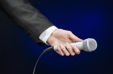 Business and speech topic: Man in black suit holding a microphone on a gray dark blue isolated background in studio