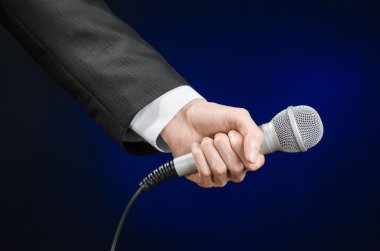 Business and speech topic: Man in black suit holding a microphone on a gray dark blue isolated background in studio