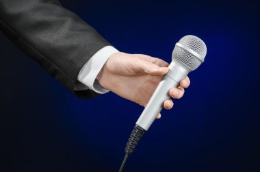 Business and speech topic: Man in black suit holding a microphone on a gray dark blue isolated background in studio