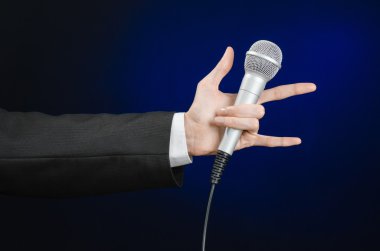 Business and speech topic: Man in black suit holding a microphone on a gray dark blue isolated background in studio