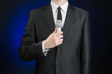 Business and speech topic: Man in black suit holding a microphone on a gray dark blue isolated background in studio