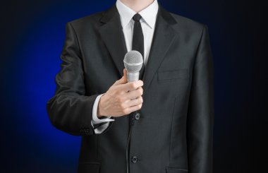 Business and speech topic: Man in black suit holding a microphone on a gray dark blue isolated background in studio